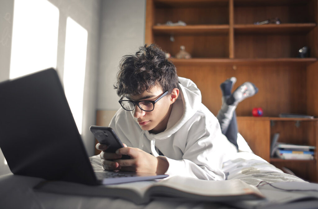 a young adult sits on their bed with their feet in the air and looks at their phone and laptop