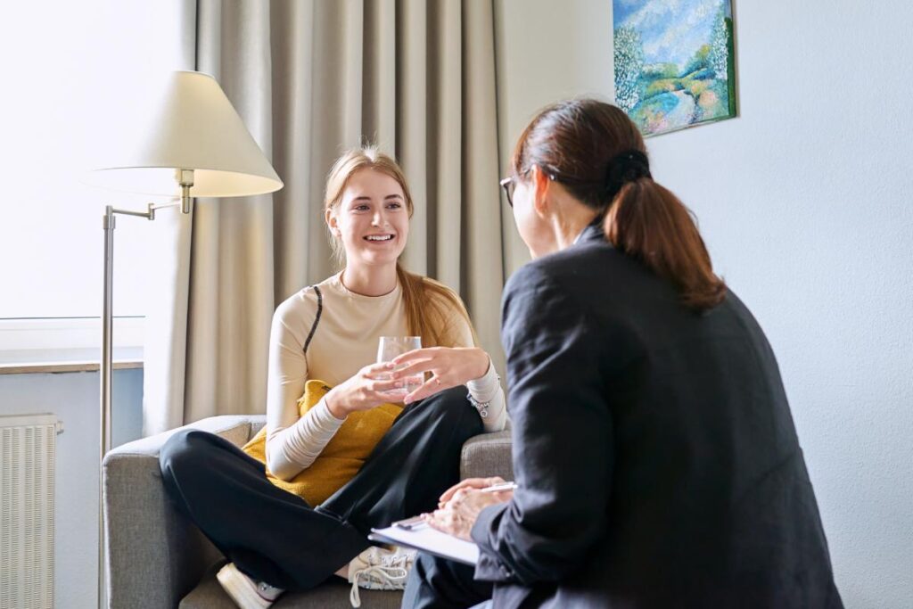 a young person sits in a chair across from a therapist while holding a glass of water