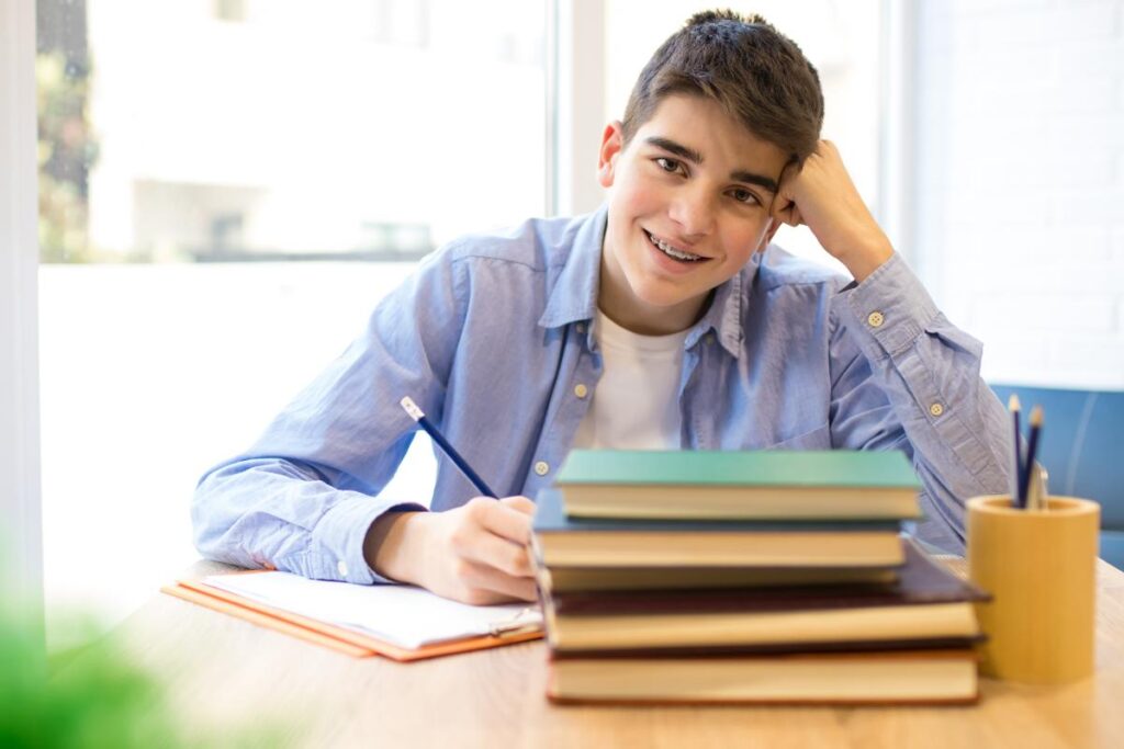 a young person with braces sits at a table in front of books and smiles at the camera while writing down in a journal for anxiety