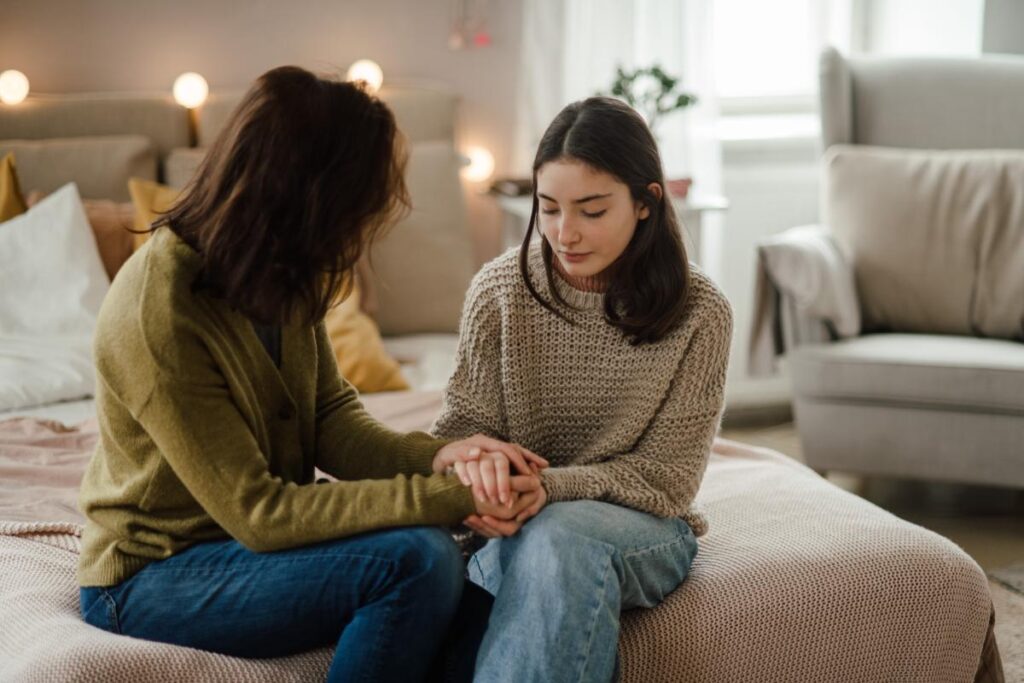 a young person holds hands with their parent while they sit on a bed and talk about mental health treatment