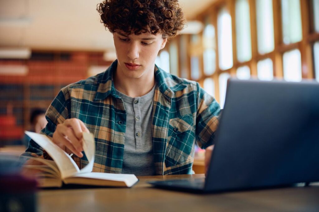 a young person sits in a library and looks at a book and laptop on a desk during mental health treatment