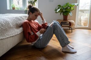 a young person sits on the floor by their bed and looks at their phone to find trauma help