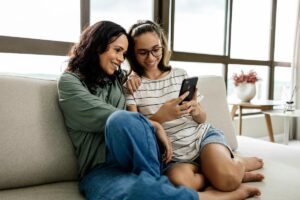 a young person and their parent look at a phone while sitting on a couch and smile learning about trauma help