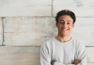 A person leans against a wood wall and smiles with arms folded